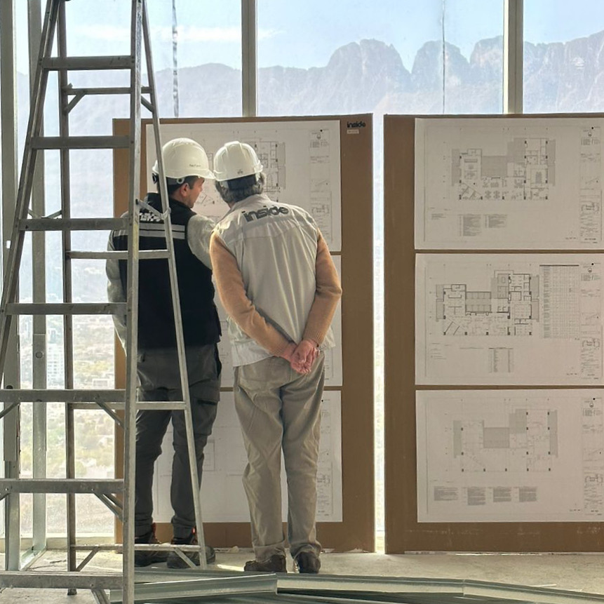 Dos integrantes del equipo de Inside, con casco, revisan los planos de un proyecto en obra frente a un ventanal con vista a la montaña